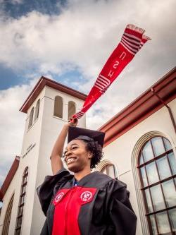 Student in graduation gown waving sash in front of the School of Communication and Media